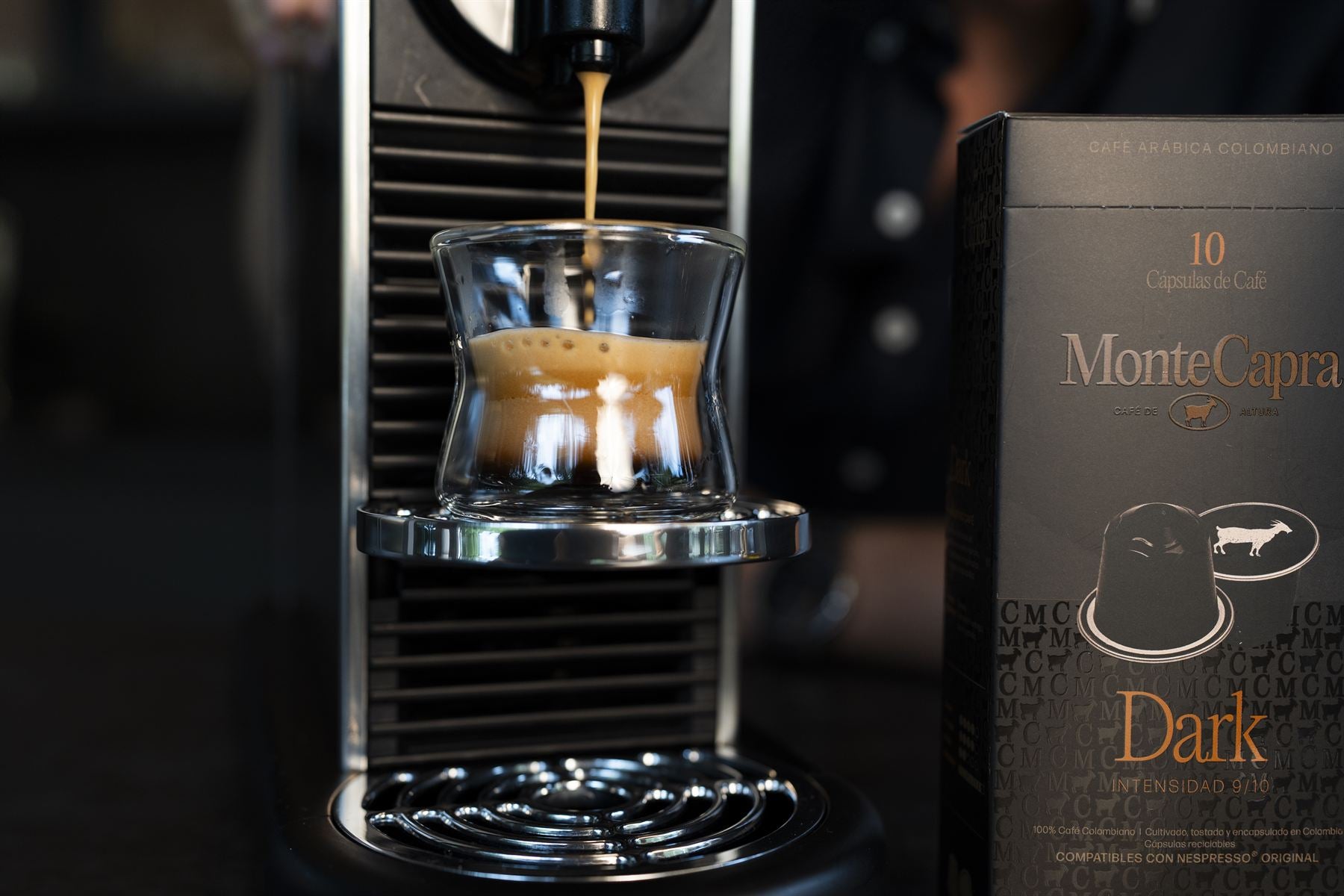 Espresso cayendo en una taza de vidrio junto a una caja de cápsulas Monte Capra Dark